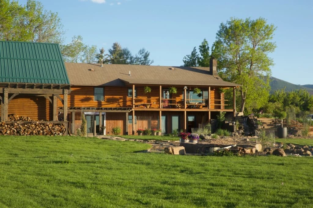 Elk Haven luxury log cabin exterior in summer with green lawn, stacked firewood, rock garden water feature, and mountain views at Big Vista Ranch Bozeman