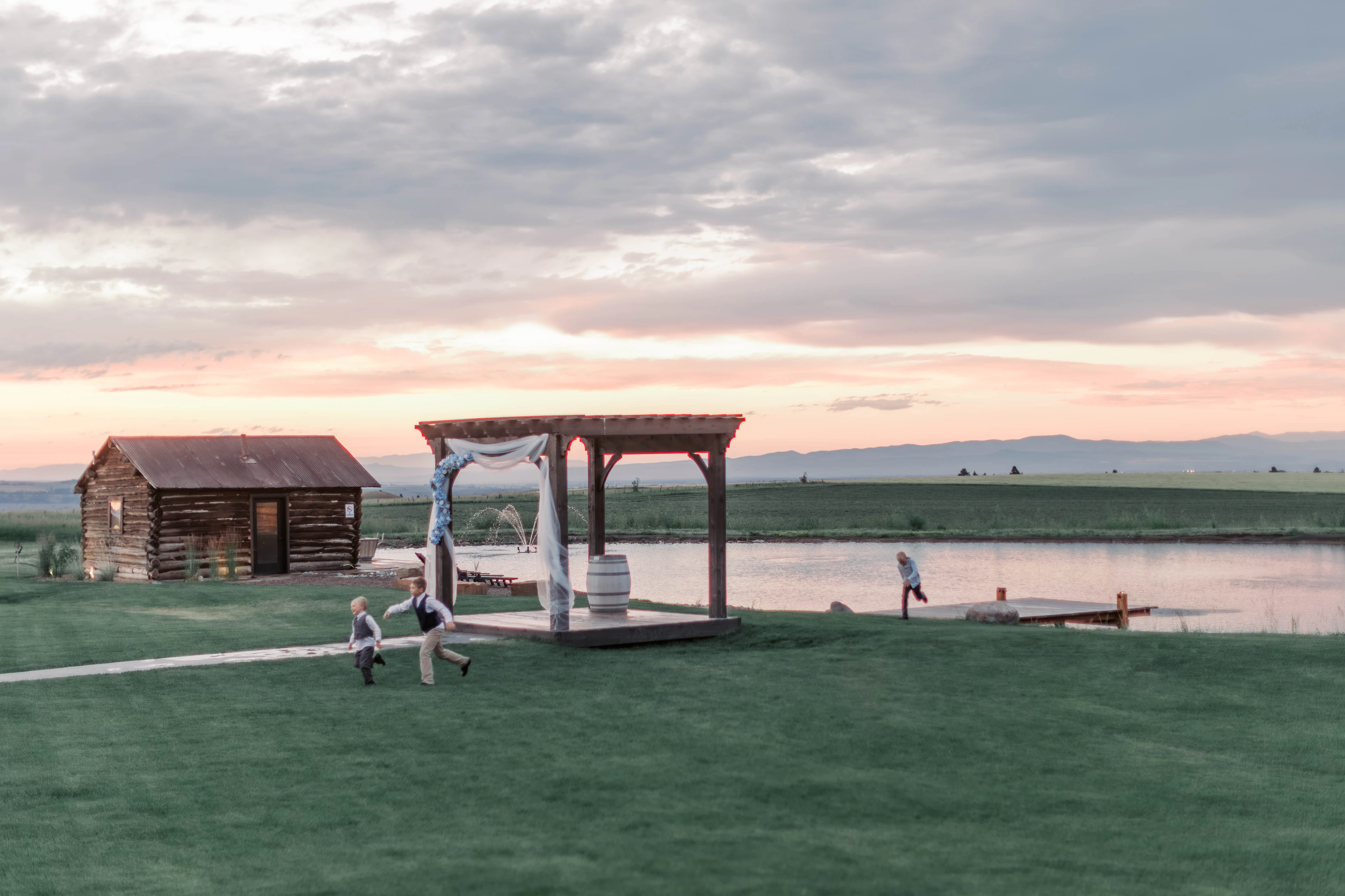 Sunset over the lakeside ceremony arch, lawn, and small timber cabin.