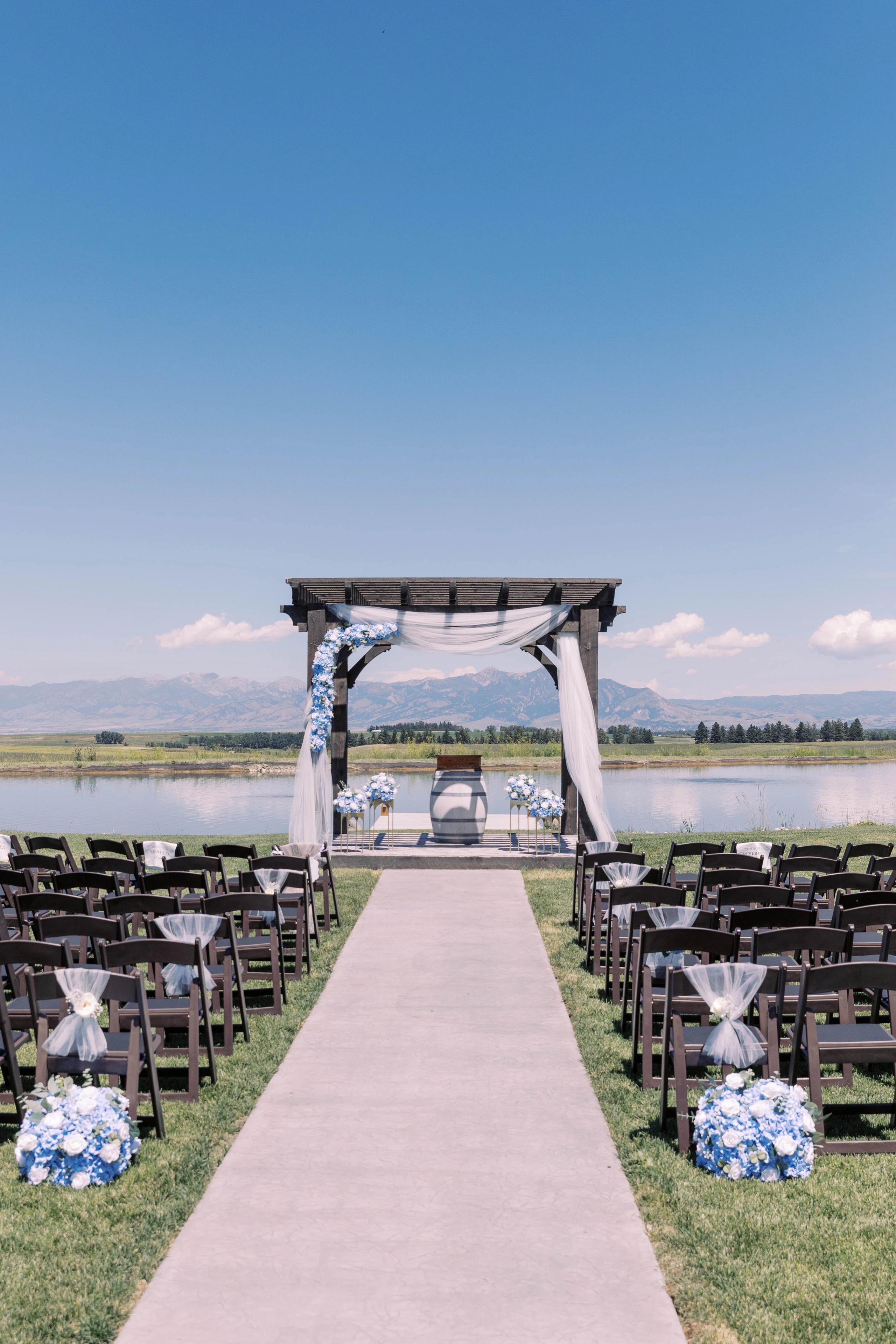 A fabric-draped wooden ceremony arch at the end of an aisle beside the lake, with mountains beyond.